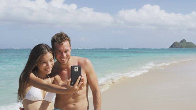 Selfie beach vacation couple taking self portrait photo with smart phone having fun using smartphone camera. Young beautiful Asian Caucasian couple on Lanikai, Oahu, Hawaii, USA.