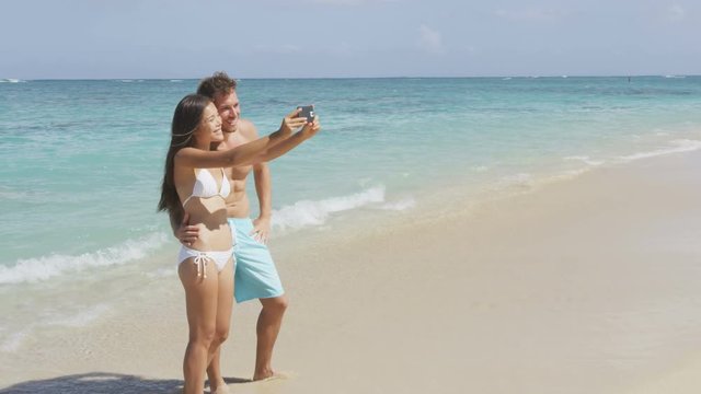 Beach vacation couple taking selfie photo with smart phone having fun using smartphone camera. Young beautiful Asian Caucasian couple taking self portrait photograph. Lanikai, Oahu, Hawaii, USA.