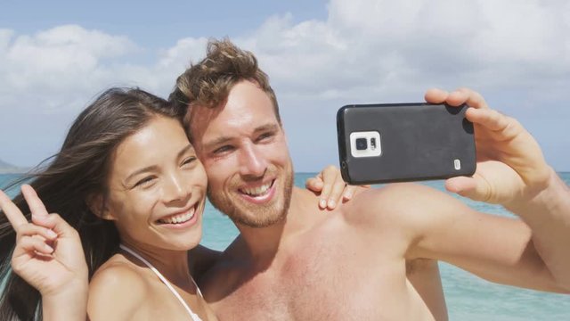 Beach holiday couple taking selfie photograph with smartphone having fun holding smart phone camera. Young beautiful multicultural Asian Caucasian couple taking self portrait photo on summer beach.