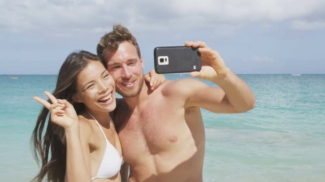 Beach vacation couple taking selfie photograph with smart phone having fun holding smartphone camera. Young beautiful multicultural Asian Caucasian couple taking self portrait photo on summer beach.