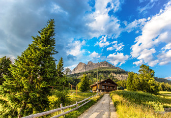 View of the mountains of the Rosengarten group (Rosengarten) with meadows and fir trees, a road and a mountain hut under a blue cloudy sky, Dolomites, Italy