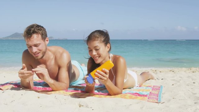 People on beach putting sunscreen suntan lotion. Sun tanning couple having fun laughing and relaxing on vacation. Adults, Asian woman and Caucasian man lying down in white sand on beautiful beach.