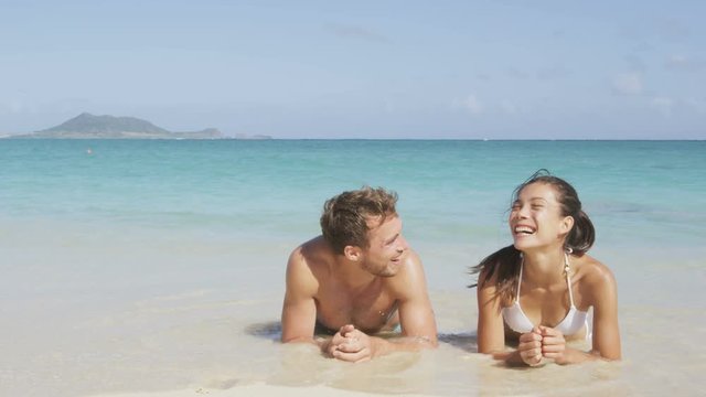 Beach couple laughing having fun with water splashing lying in sand in water edge. Woman and man on honeymoon relaxing having a good time on pristine white paradise beach. Asian woman, Caucasian man.