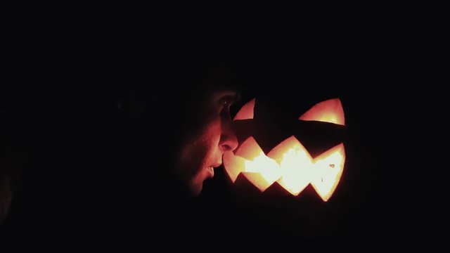 Young guy looks on burning candle inside of carved pumpkin jack o lantern face to face close up 4k