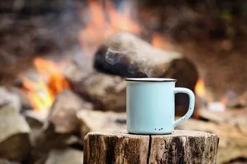 Door stickers Camping Blue enamel cup of hot steaming coffee sitting on an old log by an outdoor campfire. Extreme shallow depth of field with selective focus on mug.