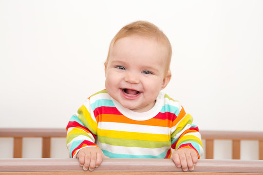 A baby is smiling in the crib and holds onto the side of the bed

