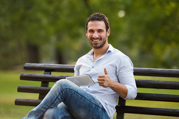 Successful businessman is showing thump up while using digital tablet and sitting at the park.
