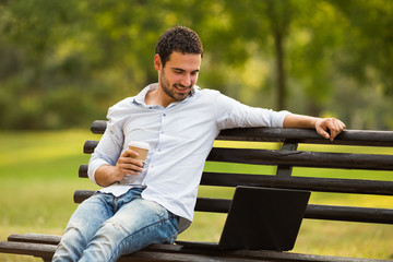 Young businessman is drinking coffee and using laptop while resting from work at the park.

