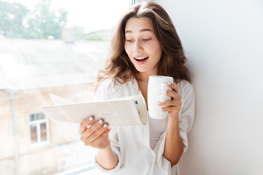 Happy young bruette woman looking at newspaper and drinking tea