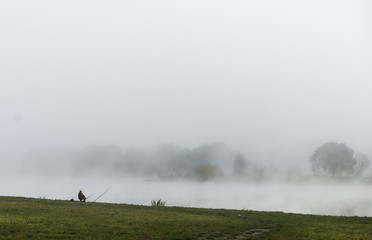 Naklejka na ściany i meble Fisher man fishing on a river bank at misty foggy sunrise