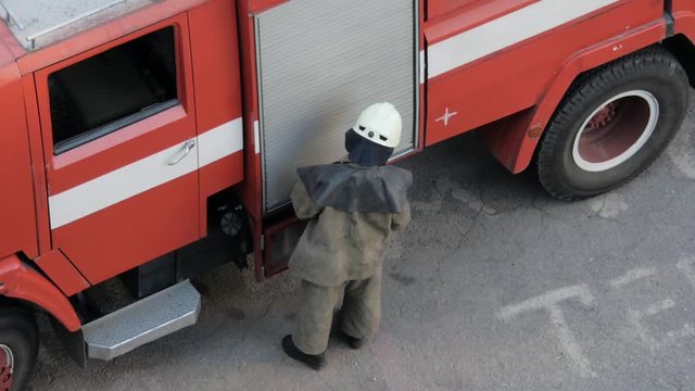 The fireman/firefighter closes the door of a fire truck and takes off the helmet. The fireman near the fire engine.
