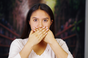 Young brunette woman wearing white sweater covering mouth using hands, facing camera, hostage...