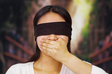 Young brunette woman wearing white sweater, blindfolded with black textile, covering mouth using...