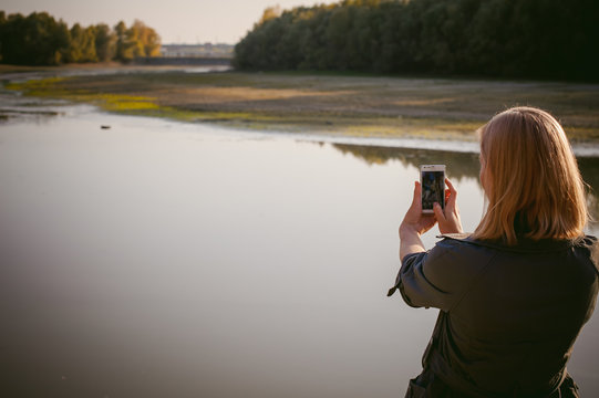 pretty blonde girl in sneakers, nylon stockings and coat. walks and photographed on a smartphone selfi on the stone shore of the lake in the last sunshine of the departing summer.