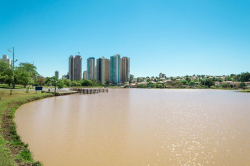 Obraz premium Lakeshore on the left with grass and some trees. Water lake and background city buildings on a sunny day.