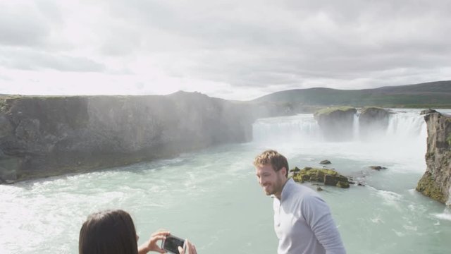 Tourist couple taking photo with smart phone having fun by waterfall Godafoss on Iceland. Girlfriend taking picture with smartphone camera of boyfriend on travel visiting  Icelandic nature attractions