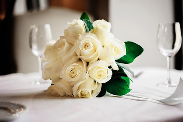 свадебный букет из белых роз лежит на белом столике в ресторане, а рядом стоят два бокала