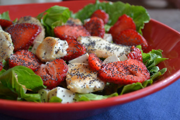 A bowl of salad with slices of roast turkey, fresh strawberry,pieces of feta cheese, green salad and poppy seeds. Summer dish in the red plate is on the navy blue cloth napkin. Healthy idea.