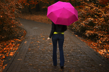 Young beautiful girl in the rain goes with the pink umbrella along the alley in the park in autumn