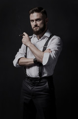 Fototapeta na wymiar Young, stylish and attractive bearded man smoking a cigar in a dark room. He is wearing in a white shirt and suspenders. He's a smart, educated hooligan and macho