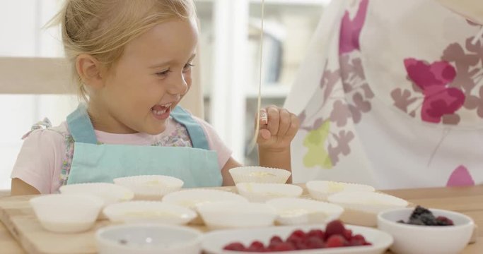 Happy little girl with empty muffin holders on table as parent picks up spoonful of batter from bowl