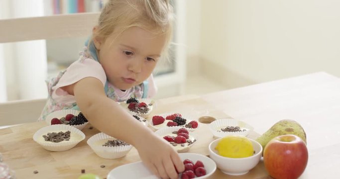 Adorable little blond girl with blond hair carefully putting berries in muffin cups ready to be baked