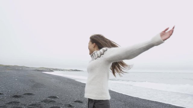 Happy free woman on Iceland in Icelandic sweater on black sand beach. Joyful girl happy swirling around smiling outdoors in nature laughing having fun. Asian Caucasian female model. RED EPIC 90 FPS.