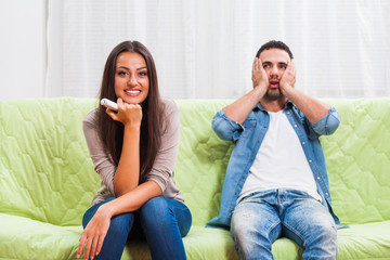 Young couple is sitting on sofa at home. Man is frustrated because he does not like what his woman is watching on TV.