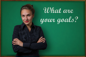 Beautiful young woman teacher (student, business woman) in classical dress standing near a blackboard with the inscription what are your goals