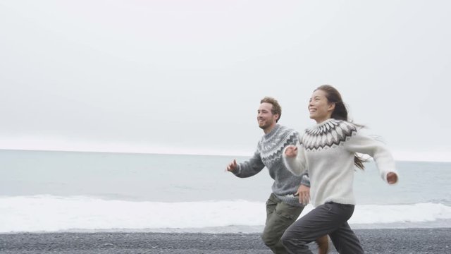 Couple having fun running on Iceland beach laughing joyful and playful wearing Icelandic sweaters on black sand beach. Woman and man model enjoying nature landscape at ocean sea. RED EPIC  90 FPS.