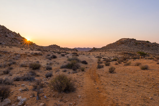 Colorful sunset over the Namib desert, Aus, Namibia, Africa. Sun star at the horizon, footpath crossing rocky desert.