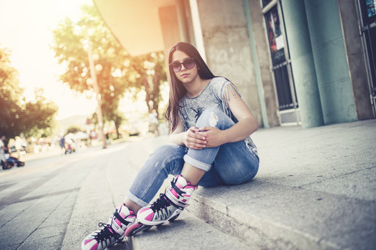 Urban vintage portrait of beautiful and attractive girl with sunglasses and roller skates. Warm summer colors and haze. Strong back light. Girl sitting on city square and enjoying hot afternoon.