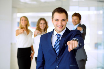 Smiling businessman  in office with colleagues in the background. Pointing by finger into the camera