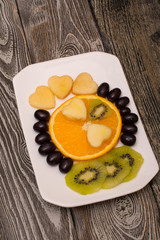 slice of orange, black grapes, kiwi and apple in the shape of a heart on a white plate, wooden background