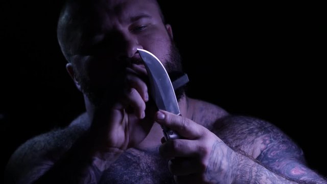 Bald tattoed bearded muscular gangster with a knife and sharpener. 4K UHD