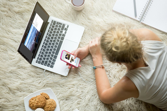 Beautiful young woman lying on white carpet with cup of tea and cookies, browsing social media, looking at pictures on smartphone and laptop, studying and relaxing at home. Top view image