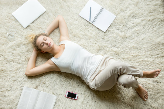 Beautiful young woman holding hands behind head while lying on white carpet on the floor in living room and sleeping after working on laptop or studying at home. Top view full length image