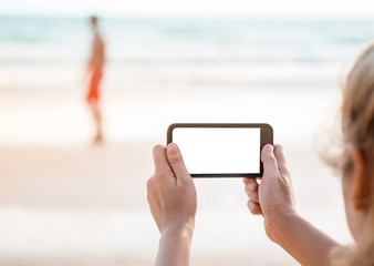 asian woman hands holding white blank screen mobile phone, take photograph of walking man with orange shorts on the beach, selective focus