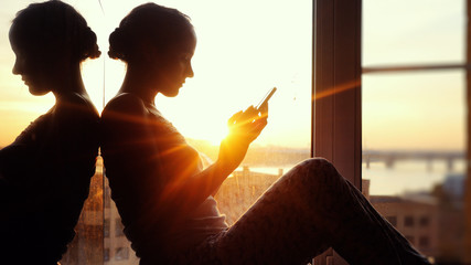 Young woman sitting on window uses smart phone  background of the sunset and city
