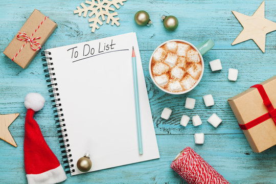 Cup of hot cocoa or chocolate with marshmallow, holiday decorations and notebook with to do list on turquoise vintage table from above, christmas planning concept. Flat lay style.