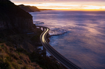 Sea Cliff Bridge on sunrise with moving traffic and dramatic beautiful sky and ocean shore on the background. The Bridge is part of NSW Grand Pacific scenic route