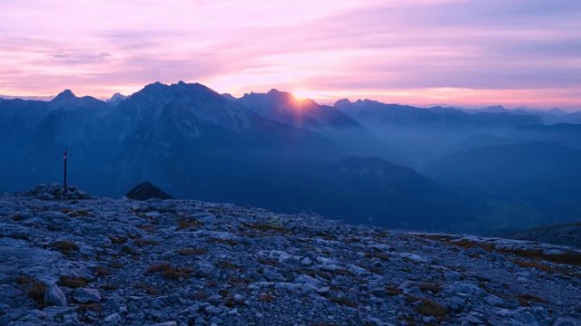 Timelapse of sunset over mountain peaks. Sun goes down over sharp peaks of Alps. Pink purple sky. Colorful flare in camera lens.