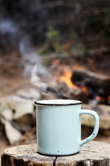 Fototapeta na wymiar Blue enamel cup of hot steaming coffee sitting on an old log by an outdoor campfire. Extreme shallow depth of field with selective focus on mug.