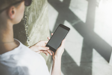 Close-up of smartphone with blank screen in female hands.Girl in white t-shirt and sunglasses standing and using gadget.In the background is a wall and a floor made of concrete.Blurred foreground.