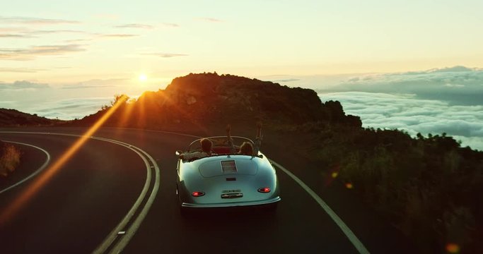 Happy couple driving on country road into the sunset in classic vintage sports car