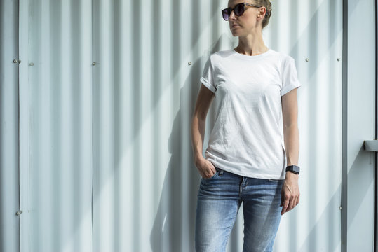 Front view, woman, dressed in a white T-shirt and blue jeans standing against a white wall. On the face of a young woman with sunglasses, on the arm of digital gadget - smartwatch. Mock up.