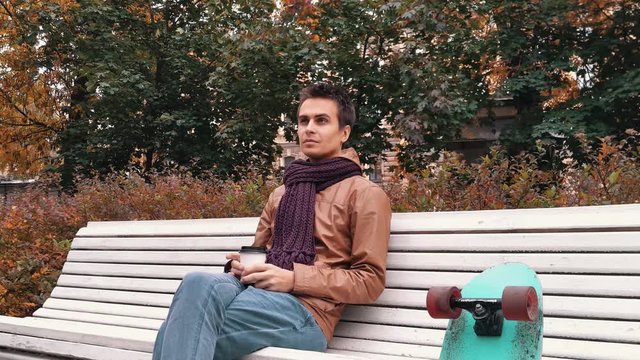 Young man with knitted scarf with skateboard sitting on bench in autumn park drinks coffee out of paper cup