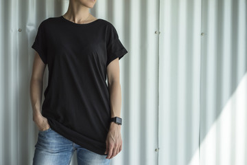 Girl in the black T-shirt and blue jeans standing against a white wall with his hand in his pocket. On hand of the girl digital gadget-smartwatch.Mock up.