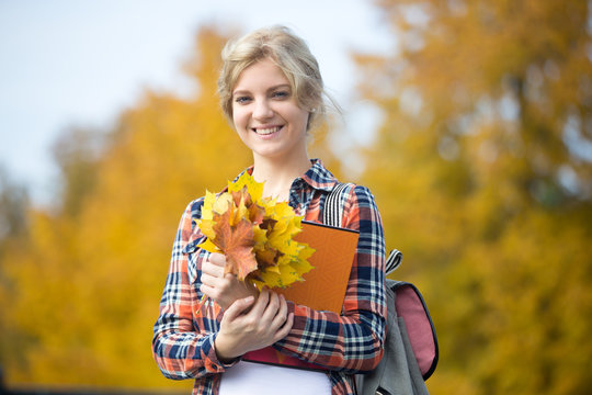 Portrait of smiling attractive female young student outdoors holding yellow maple leaves and copybooks, yellow background. Fall time. Back to school concept photo, looking at the camera