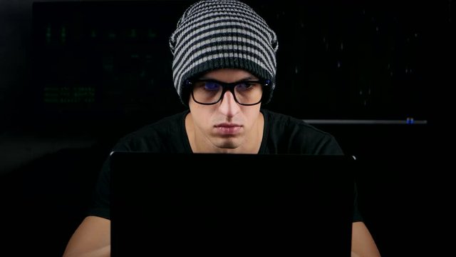 Male hacker with glasses and hat working on a computer in a dark office room